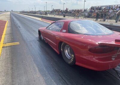 Red car racing on drag strip with spectators.